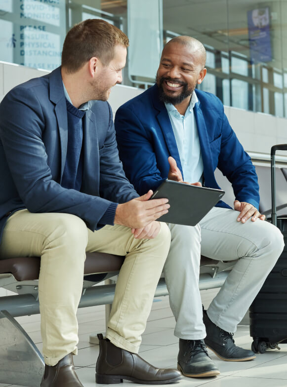 two men sitting in the airport with an ipad and a suitcase