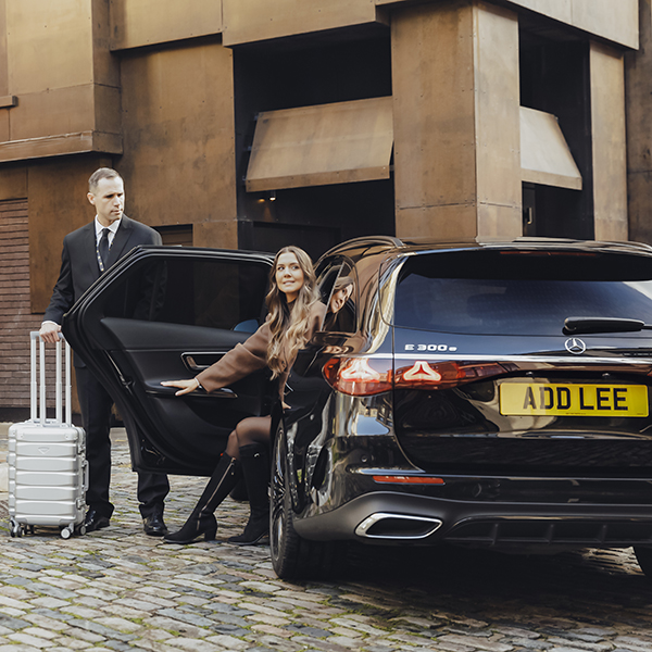 Woman sitting in an Addison Lee taxi while a chauffeur opens the door for her with her luggage