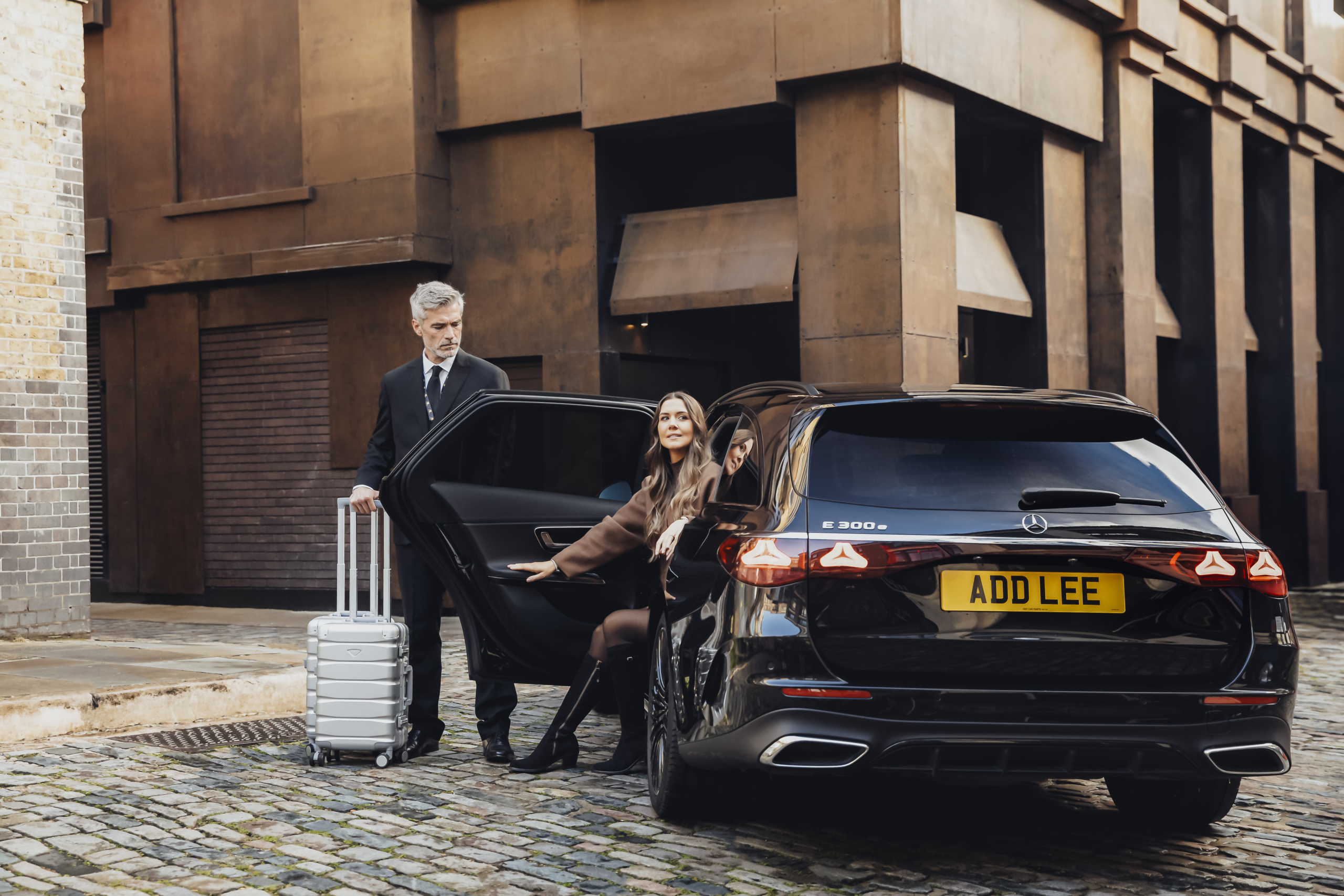 Woman getting out of an executive car with an Addison Lee driver holding the door open for her and helping with her luggage